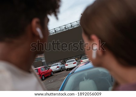 Young multiracial couple watching movie on autocinema screen while standing together on car parking, selective focus. Free time, leisure and entertainment concept