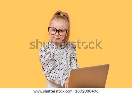 Blonde smart girl learn online using a laptop. Large black-rimmed glasses. Yellow isolated background.