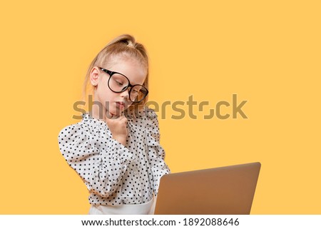 Cute blonde girl looks at her laptop and thinks. Large glasses for vision. Yellow isolated background.