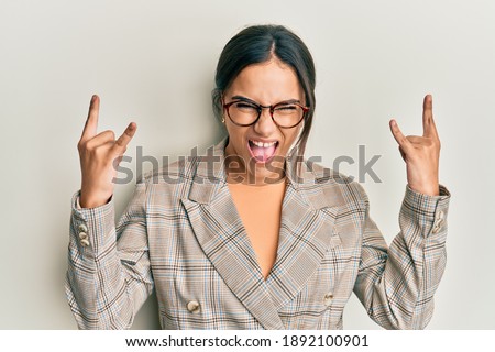 Young brunette woman wearing business jacket and glasses shouting with crazy expression doing rock symbol with hands up. music star. heavy music concept. 