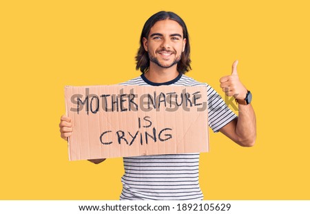 Young handsome man holding mother nature is crying protest cardboard banner smiling happy and positive, thumb up doing excellent and approval sign 