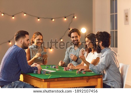 Cheerful friendly company of men and women sitting at a poker table and enjoying the game. Friends play a card game at home or in a club. Evening entertainment concept. Royalty-Free Stock Photo #1892107456