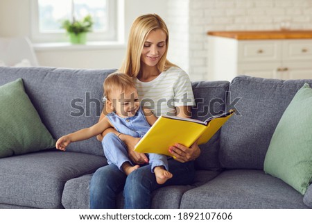 Happy mother sitting on sofa with her baby on knees and reading book or watching album with pictures together at home with room interior at background. Happy childhood and motherhood concept