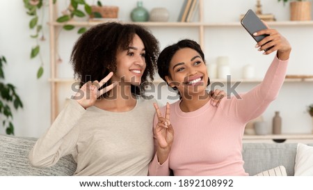 Beautiful women take photos on phone at home party. Smiling young african american friends make sign of peace with hand and do selfie for modern blog or social networks in living room, panorama