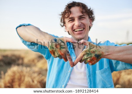 Close-up picture of male artist showing heart sign with dirty with paint hands on wheat field in summer.Painting workshop in rural countryside. Artistic education concept.Outdoors leisure activities.