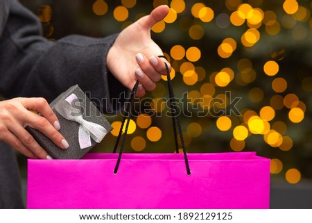 Woman takes out a silver gift box from a shopping bag on a blurred lights background. Empty space
