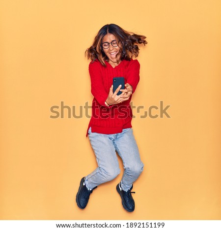 Young beautiful latin woman wearing glasses smiling happy. Jumping with smile on face make selfie by smartphone over isolated yellow background