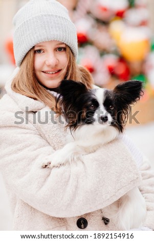 Beautiful teenage girl in a gray hat with dog Papillon in her arms on the winter Christmas streets. backstage new year photo shoot with a small pet. 