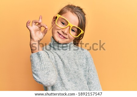 Little beautiful girl wearing casual clothes and yellow glasses doing ok sign with fingers, smiling friendly gesturing excellent symbol 