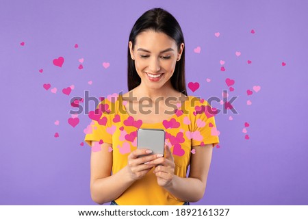 Love Mail Concept. Smiling young woman using smartphone, receiving love text message with pink hearts flying away from gadget screen isolated on purple studio background. Dating App. Royalty-Free Stock Photo #1892161327