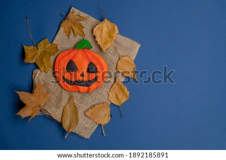 Jack O'Lantern gingerbread cookie lies on sackcloth (or burlap) by fallen birch leaves on the blue background. Smiling pumpkin face. Halloween homemade baking theme. Copy space for your text.