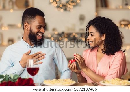 Will You Marry Me. Smiling african american woman proposing to her boyfriend, holding and showing open box, offering marriage ring to happy man, getting engaged in restaurant during romantic date Royalty-Free Stock Photo #1892187406