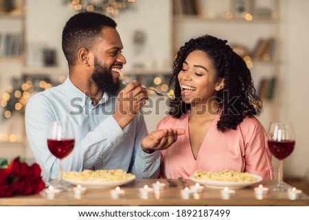Affectionate young black couple having dinner, guy feeding his girlfriend with pasta. Cute lovers having romantic date, flirting, having fun, celebrating St Valentine's Day or holiday Royalty-Free Stock Photo #1892187499