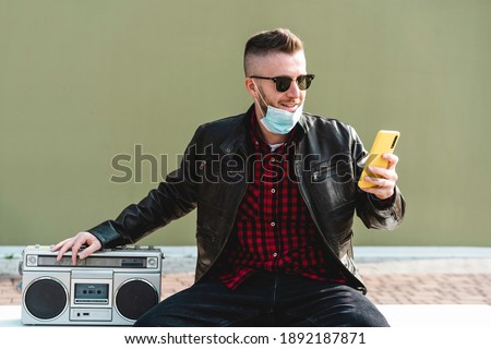 Man video calling against green wall background while listening music with a retro boombox - Cool man wearing open face protective mask looking phone camera for live broadcast during covid 19 time