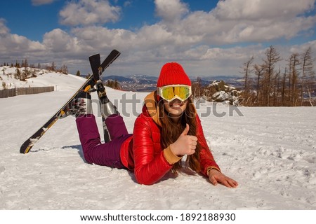A teenage girl in a red jacket and ski glasses lies in the snow. ski-skier's photo shoot. ideas photo in the snow