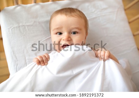 Closeup of adorable smiling blonde baby laying in wooden crib under blanket. Happy little kid infant woke up after sleeping, playing with duvet and looking at camera, lying in baby bed