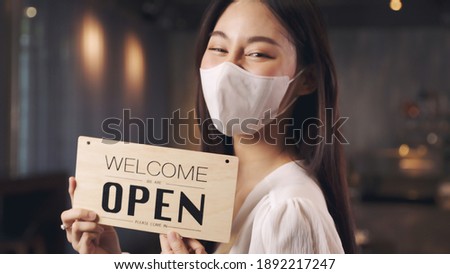 Business owner attractive young Asian woman in apron hanging we're open sign on front door  welcoming clients to new cafe. Happy waitess with protective face mask holding open sign at cafe .