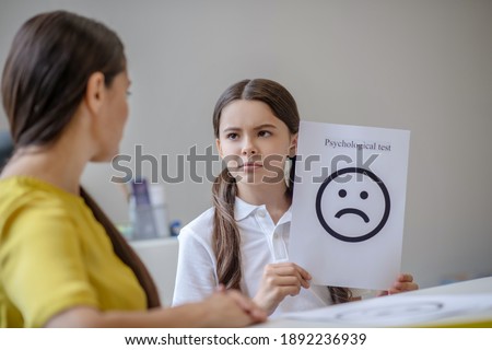 Psychological test. Sad school-age girl holding picture with painted sad emotion and woman psychologist opposite