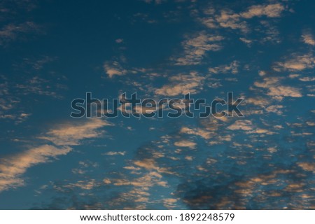 A cloudy landscape, in a bright blue sky, a series of small white clouds with a golden tint of the setting sun is gradually being covered by a dark cloudy haze.