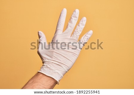 Hand of caucasian young man with medical glove over isolated yellow background counting number 5 showing five fingers