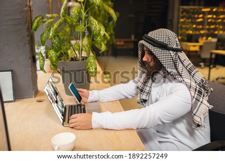Conference. Arabian businessman working in office, business centre using device, gadget. Modern saudi lifestyle. Man in traditional wear and scarf looks confident, busy, handsome. Ethnicity, finance.