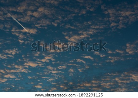 In a skyscape, a contrail cuts through the bright blue of the winter sky and cirrus clouds tinged with the golden hues of the setting sun.