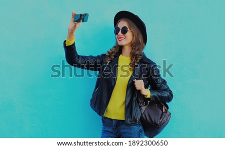 Stylish woman taking selfie picture by smartphone wearing black hat, backpack on a blue background