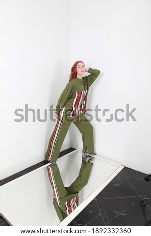Full length of female ginger model posing in trendy altheisure style pants suit, posing standing over white walls background. Copy space for text. Catalog fashion photo shoot. Image in high key.