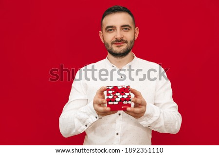 Young bearded happy European man in white shirt holding a gift box on red background. Happy valentines day. Gifts, fun, smile, love day, holiday.