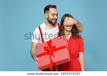 Funny young couple friends man woman in white red clothes hold present box with gift ribbon bow cover eyes with hand isolated on blue background. Valentine's Day Women's Day birthday holiday concept Royalty-Free Stock Photo #1892354533