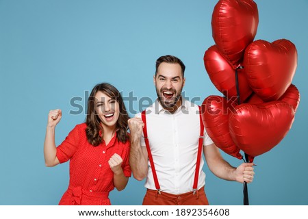 Joyful young couple friends man woman in white red clothes celebrating birthday holiday party hold bunch of air inflated helium balloons doing winner gesture say yes isolated on blue color background
