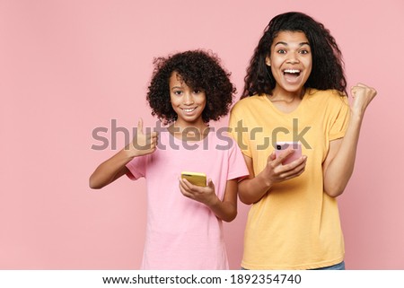 Happy african american young woman little kid girl sisters in t-shirts using mobile phone typing sms message showing thumb up winner gesture isolated on pink background studio. Family day concept