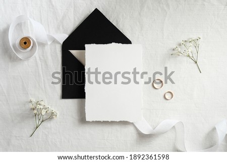 Wedding invitation card mockup. Blank white paper card, black envelope, golden rings, ribbon, flowers top view.
