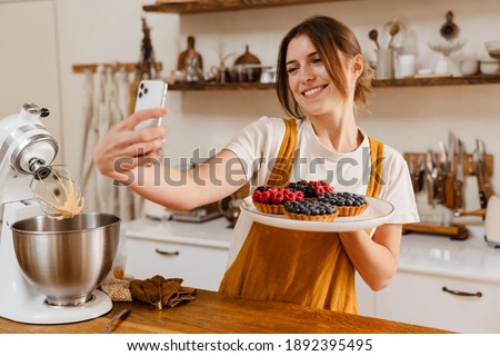 Beautiful smiling pastry chef woman taking selfie with tarts on cellphone at cozy kitchen