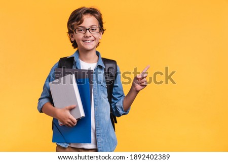 Smiling little boy pointing at copy space in casual clothes with books for studing at school isolated over yellow background Royalty-Free Stock Photo #1892402389