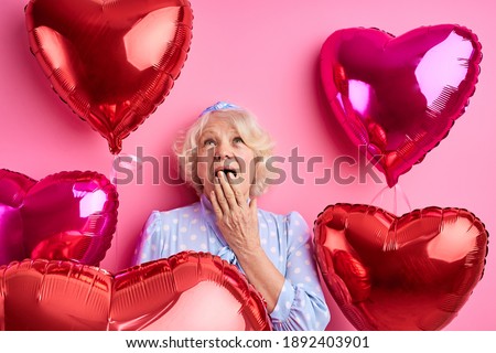 surprised elderly female in festive clothes,stand with air balloons, celebrates anniversary or Valentine's Day, enjoys celebration, poses over pink studio wall. Pensioner on party
