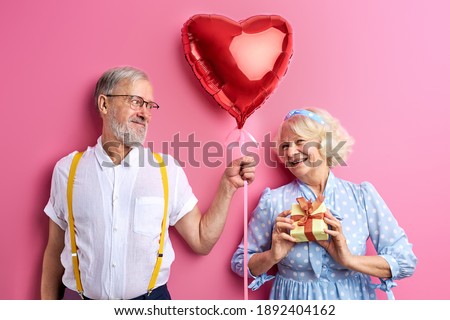 elderly man and woman please each other, celebrating holidays together, giving gifts to each other, isolated on pink backrgound. portrait
