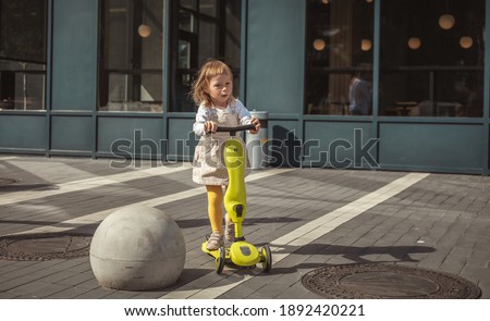 Little Caucasian girl rides a scooter in a city full of obstacles. Modern city walls with 
restaurant glass showcase. Kids safety