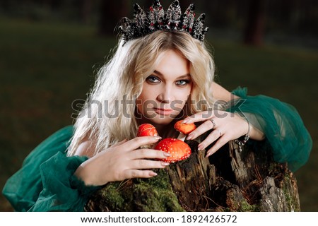 A beautiful blonde young woman in a long green dress and a diadem on her head in the forest. girl sitting near the old stump with amanitas. Solar glare. Fantasy. fairy tale.