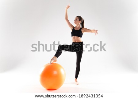 A slender girl in leggings and a sports top, stands on a white background, putting one leg on a fitness ball. Girl athlete on a white isolated background, with a ball