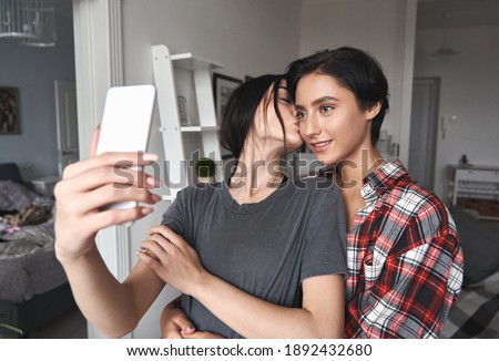 Two young happy women lesbian lgtbq couple holding smartphone kissing, hugging, using mobile phone at home, taking selfie shoot for social media application or streaming in app virtual chat together.