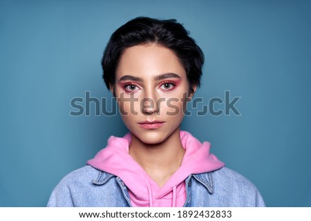 Confident stylish cool gen z teen girl, short-haired zoomer teenager with trendy pink makeup on pretty face wearing denim looking at camera isolated on blue background. Close up headshot portrait. Royalty-Free Stock Photo #1892432833