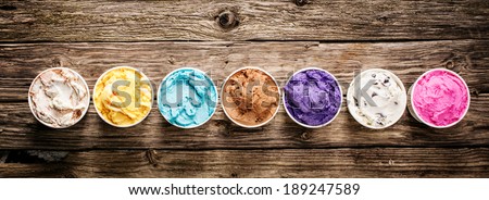 Row of assorted flavors and colors of gourmet Italian ice cream served in plastic takeaway tubs on a rustic wooden table, horizontal banner format with copyspace