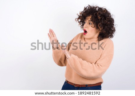 Displeased Young beautiful Arab woman wearing knitted sweater standing against white background keeps hands towards empty space and asks not come closer sees something unpleasant