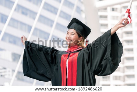 Portrait of Asian female student wearing graduated uniform and holding certification while sitting on stairs. Education Concept. Royalty-Free Stock Photo #1892486620