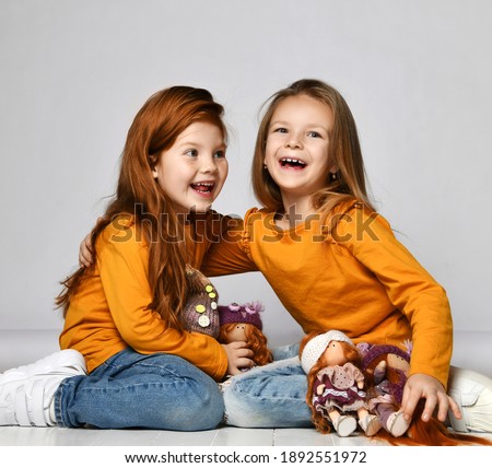 Two frolic laughing kid girls best friends in similar clothes orange sweatshirts and blue jeans sit on floor in kindergarten playing together with their favorite dolls over gray wall background