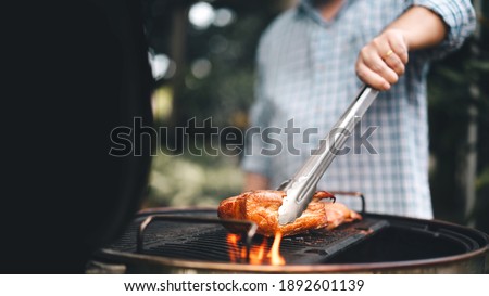 Man hand grilling barbecue on fire at backyard on day. Family dinner outdoor style bbq activity. Royalty-Free Stock Photo #1892601139