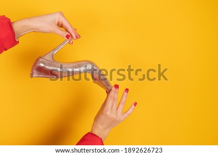 Cropped view of stylish golden shoe in female hands with perfect red manicure. Yellow studio background with copy space. Great shoes sale concept. High quality photo