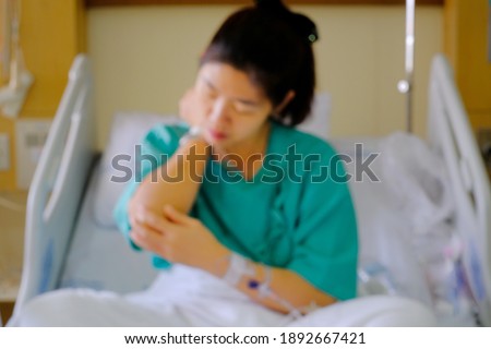 An out of focus picture of an Asian woman being treated for her illness in a hospital room, sitting on a bed with a green uniform and saline solution injected on her arm, holding her hand in pain.