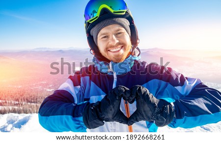 Happy male athlete skier smiles and shows heart from hands with sunlight. Winter active vacation concept.